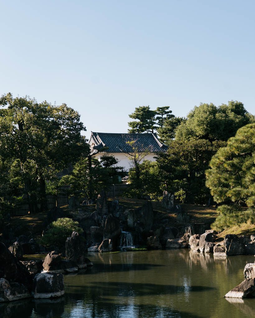 A garden pond with rocks and a small waterfall, with a white-walled, tiled-roof building among pine trees behind, in the grounds of Nijo Castle in Kyoto