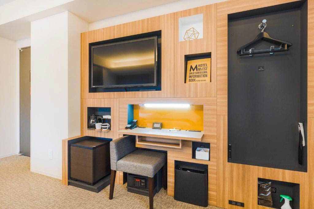 Hotel room storage wall with wall-mounted TV, work desk, chair, mini fridge, and in-room safe
