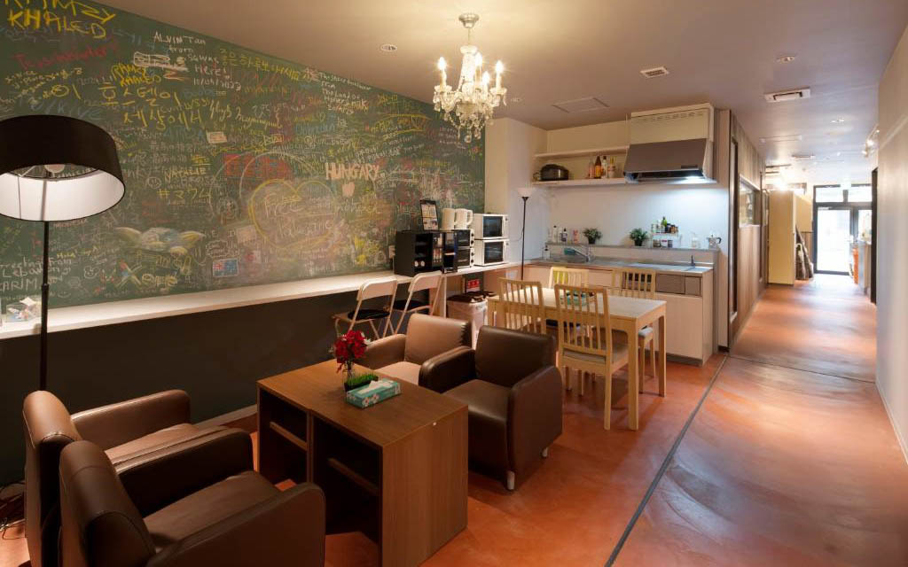 Hostel common area with a kitchen, dining table, sofas, chandelier, and a chalkboard wall covered in guest messages