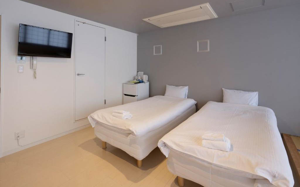Private hostel twin room with two single beds, wall-mounted TV, and mini fridge