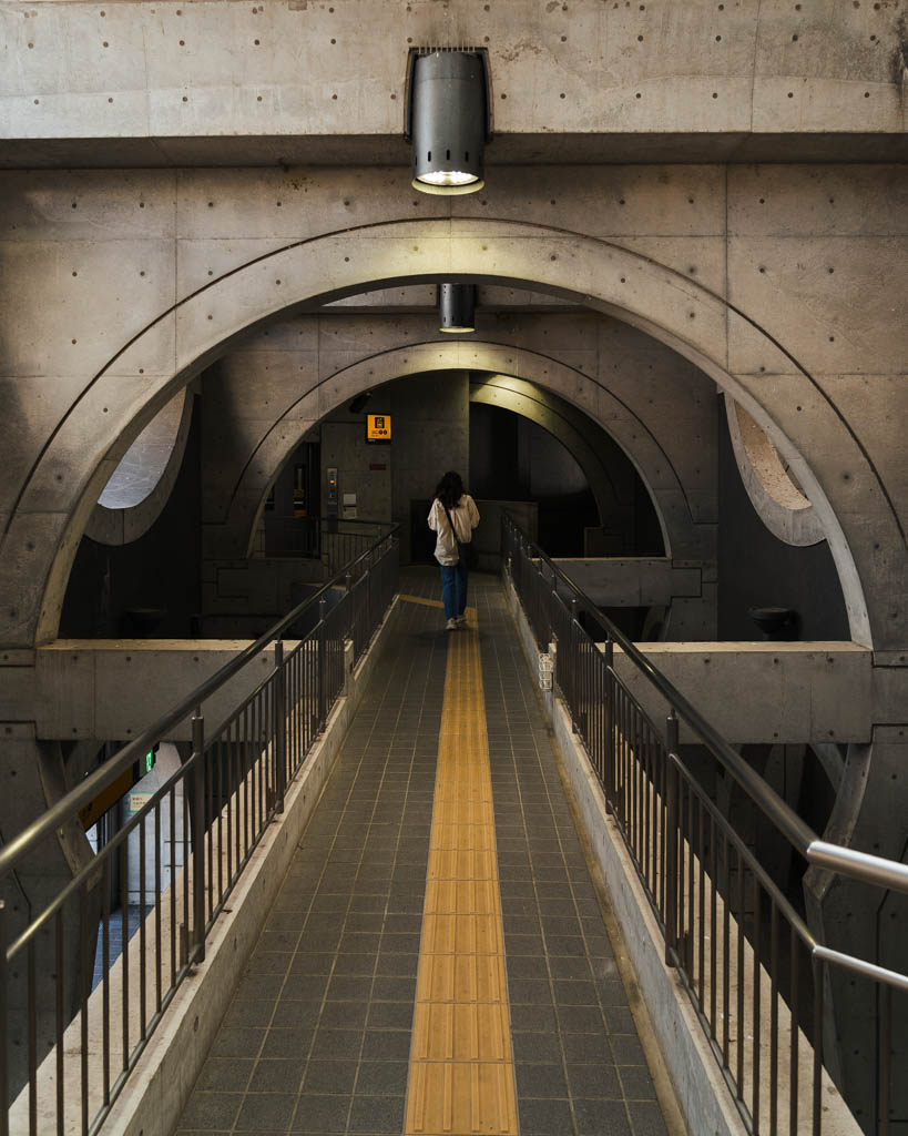 A woman walking along a walkway beneath large concrete arches inside Keihan Uji Station