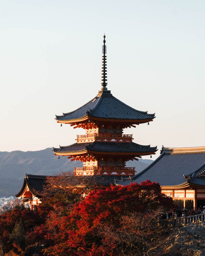 Three-storey red pagoda at Kiyomizu-dera in golden evening light, with bright red autumn maple leaves in the foreground and mountains behind