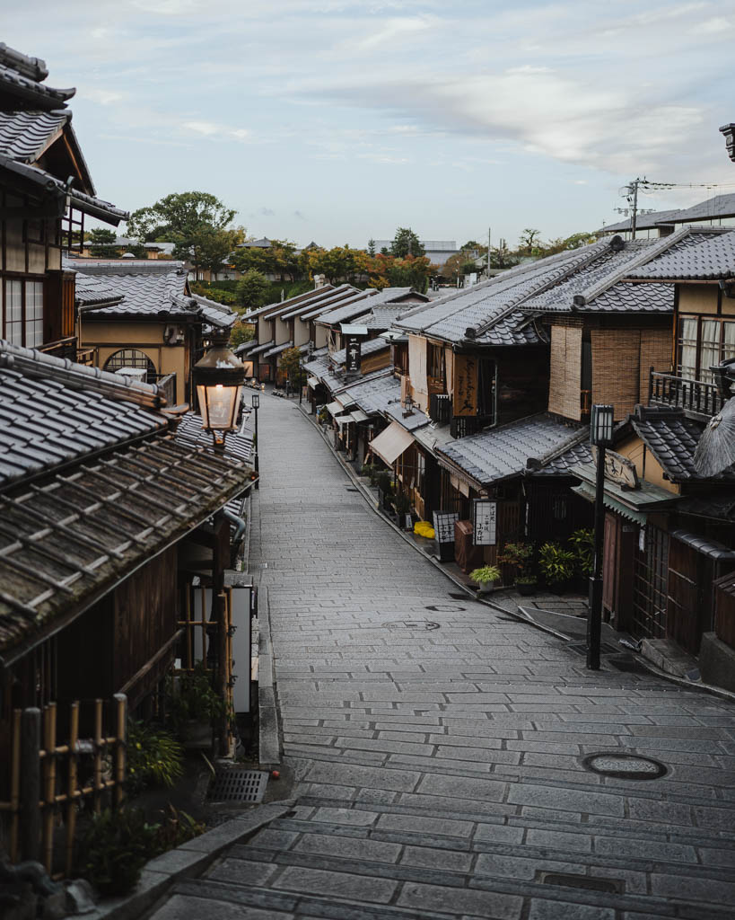 An empty stone-paved Ninenzaka street lined with traditional wooden buildings and tiled roofs in the early morning, with autumn trees in the distance