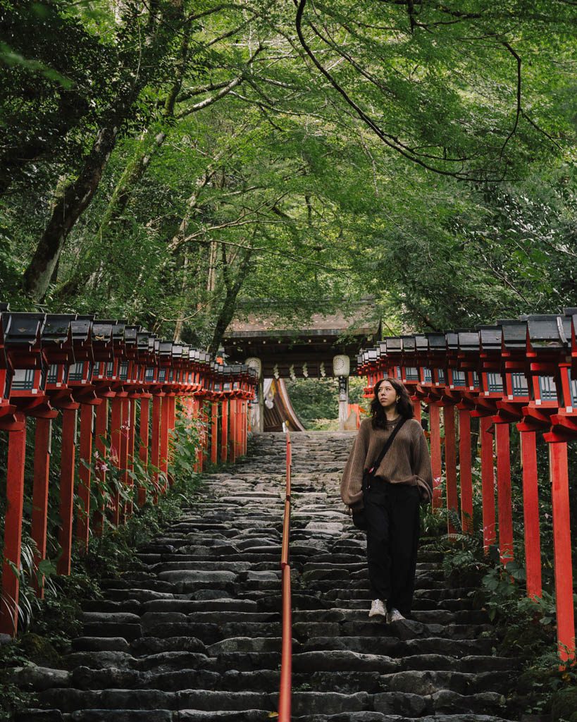 A woman walking down the stone steps of the red lantern staircase leading up to Kifune Shrine, with green maple trees arching overhead