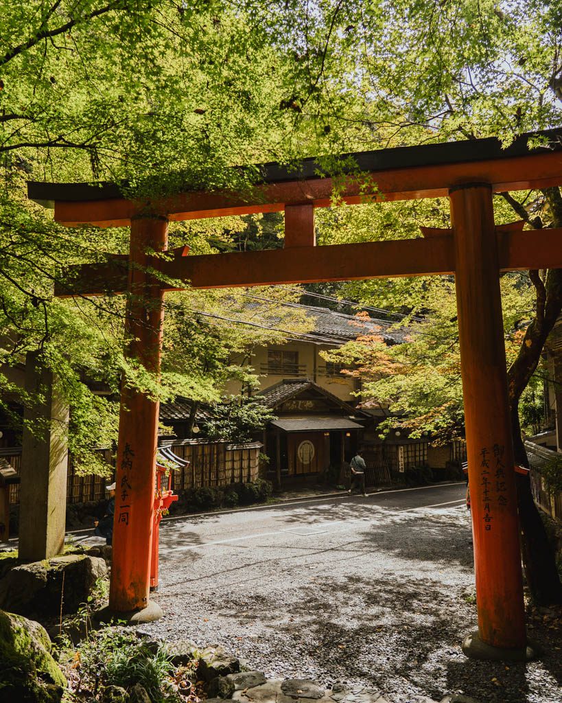A red torii gate framed by green maple leaves with the first hints of orange at Kifune, with a traditional wooden building across the road