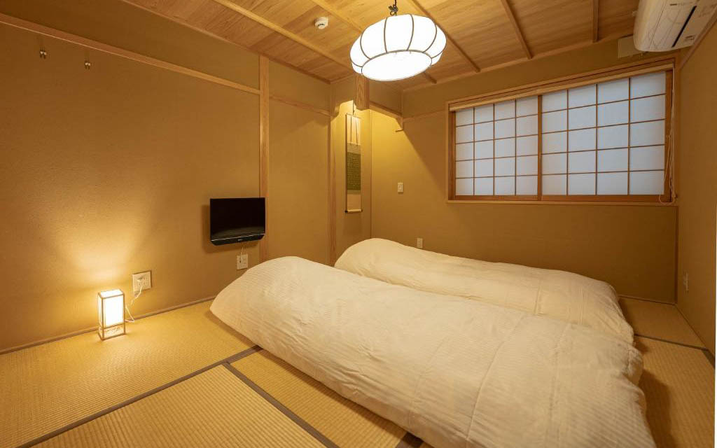 Tatami room with two futon beds laid out, paper lantern, wooden ceiling beams, and shoji window