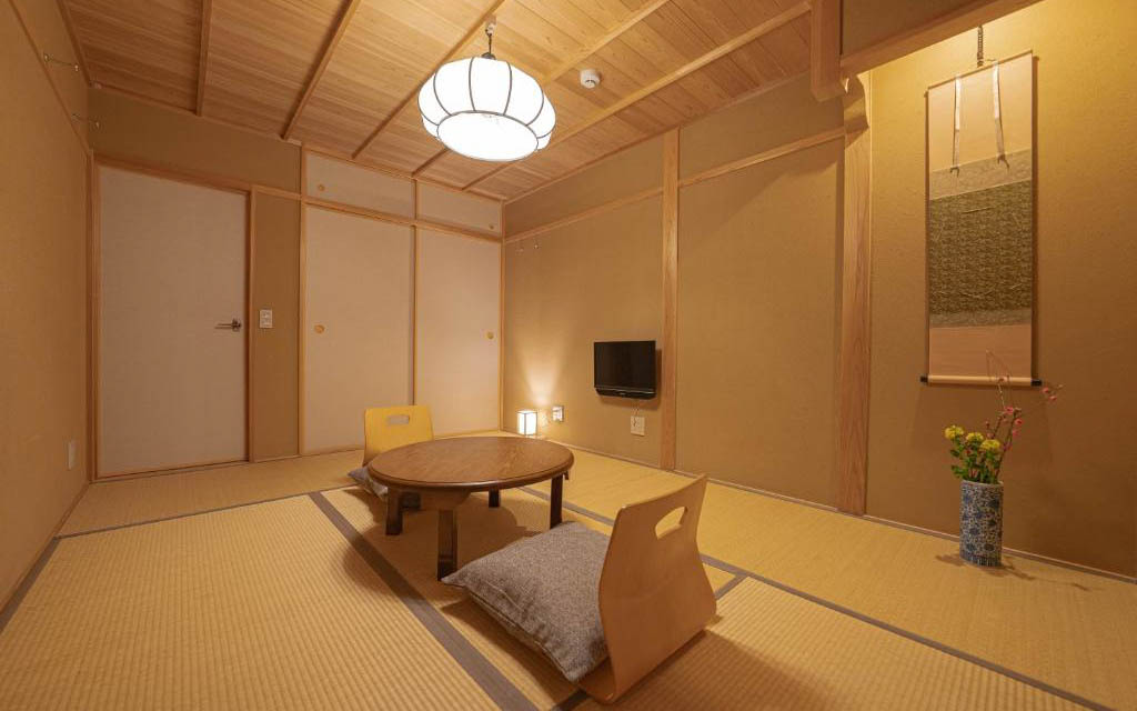 Tatami room set up with a low wooden table, floor cushions, wall-mounted TV, and a hanging scroll