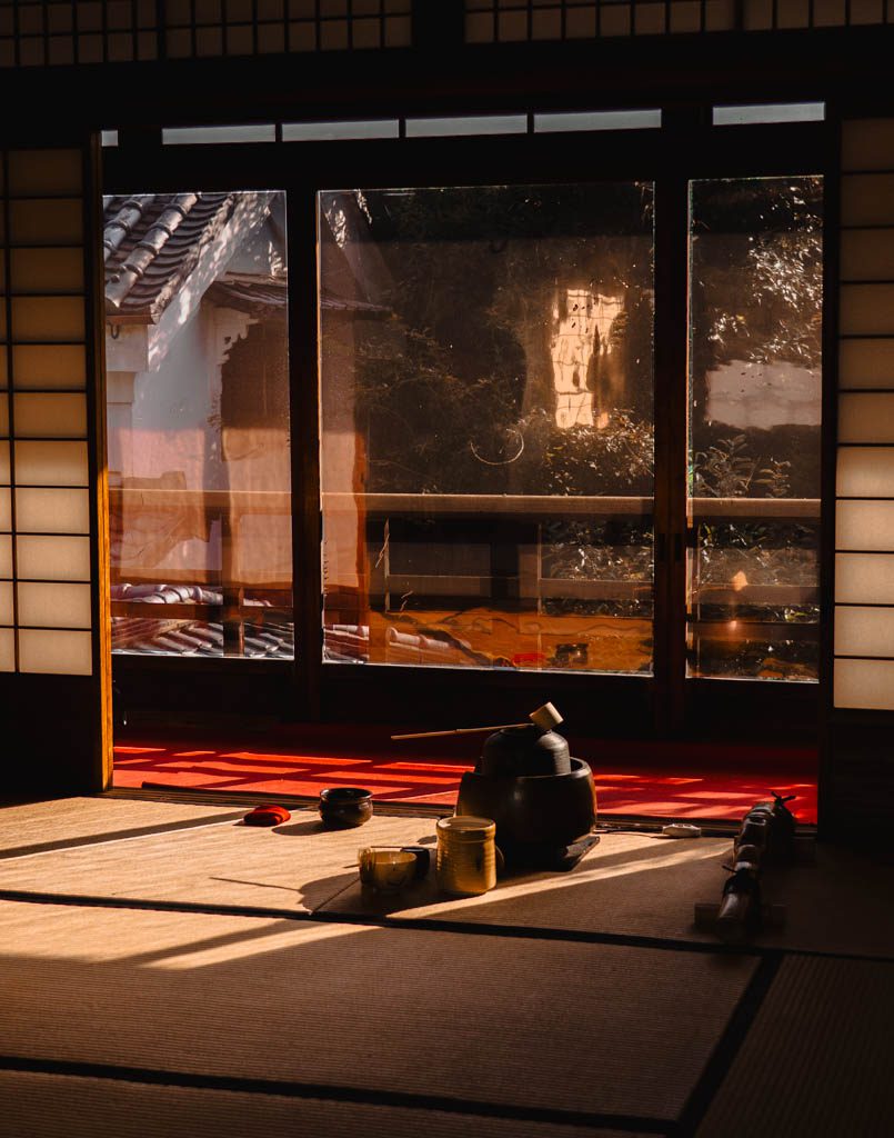 Tea ceremony utensils laid out on tatami mats in a sunlit tea house in the Gion district, with a red carpet and garden visible through glass doors