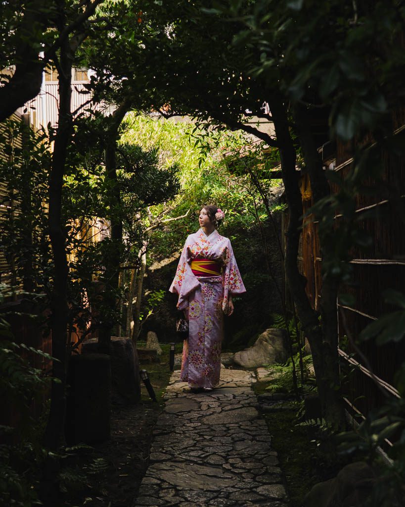 Woman in a pink floral kimono walking along a stone path through a shaded Japanese garden in Kyoto