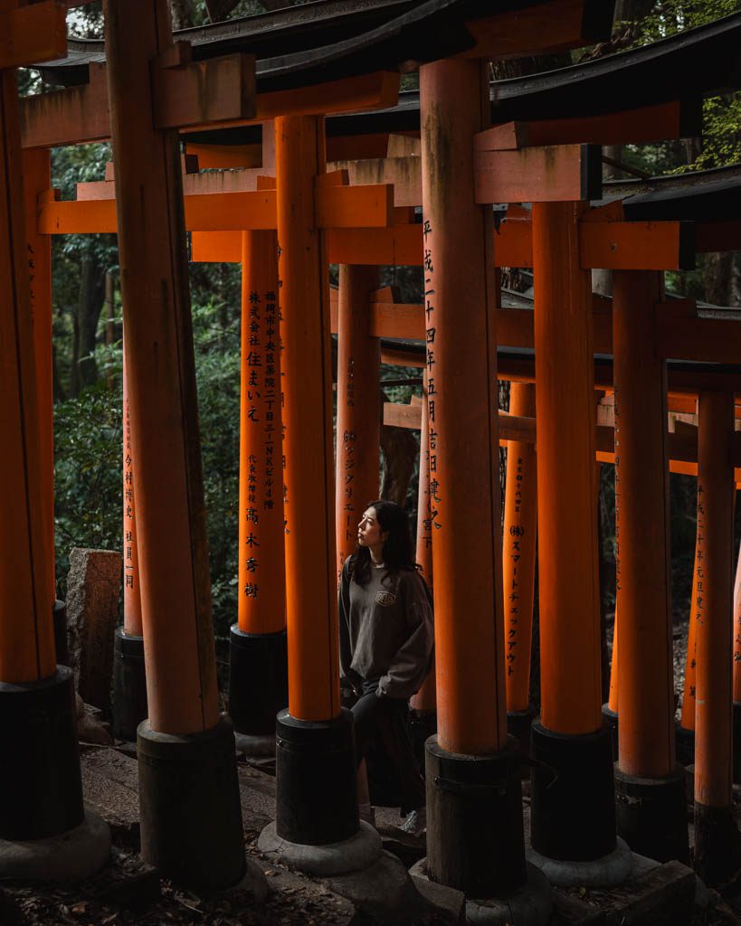 A woman looking up between rows of tall orange torii gates with black bases on the hiking path at Fushimi Inari Shrine in Kyoto