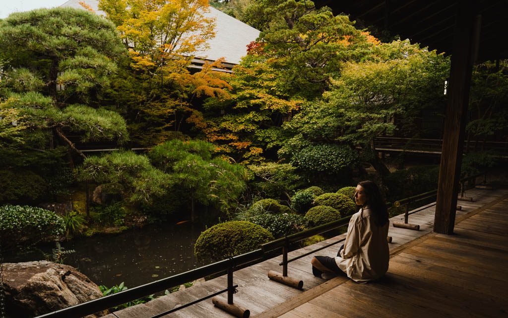 Woman sitting on a wooden veranda looking out over a pond and autumn garden at Eikan-do temple in Kyoto