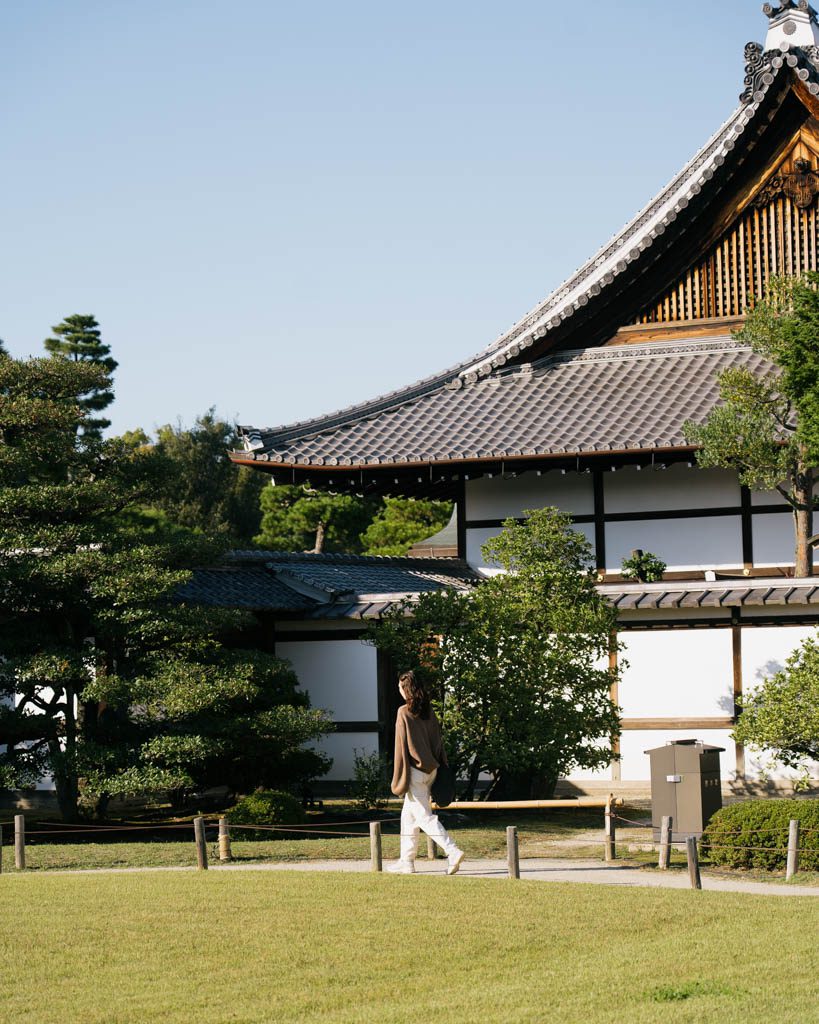 A woman walking across the lawn in front of a traditional white-walled building with a sweeping tiled roof in the grounds of Nijo Castle in Kyoto