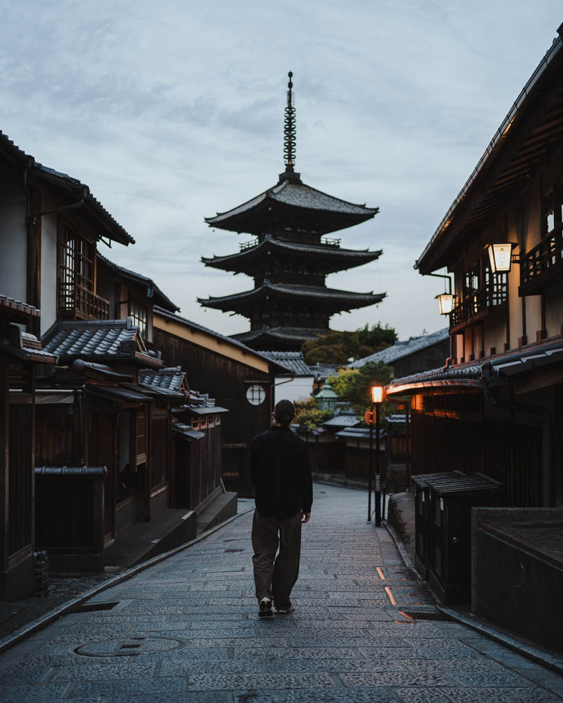A person walking down a quiet stone street towards a five-tier pagoda in the blue light before sunrise, with lanterns glowing on the wooden buildings either side